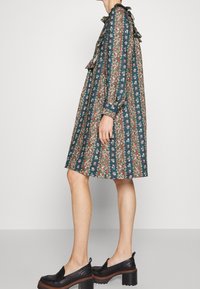 Květinové šaty s volantovým límcem a rukávy, se zelenými, červenými a bílými vzory na tmavě modrém pozadí. Součástí jsou černé platformové boty.