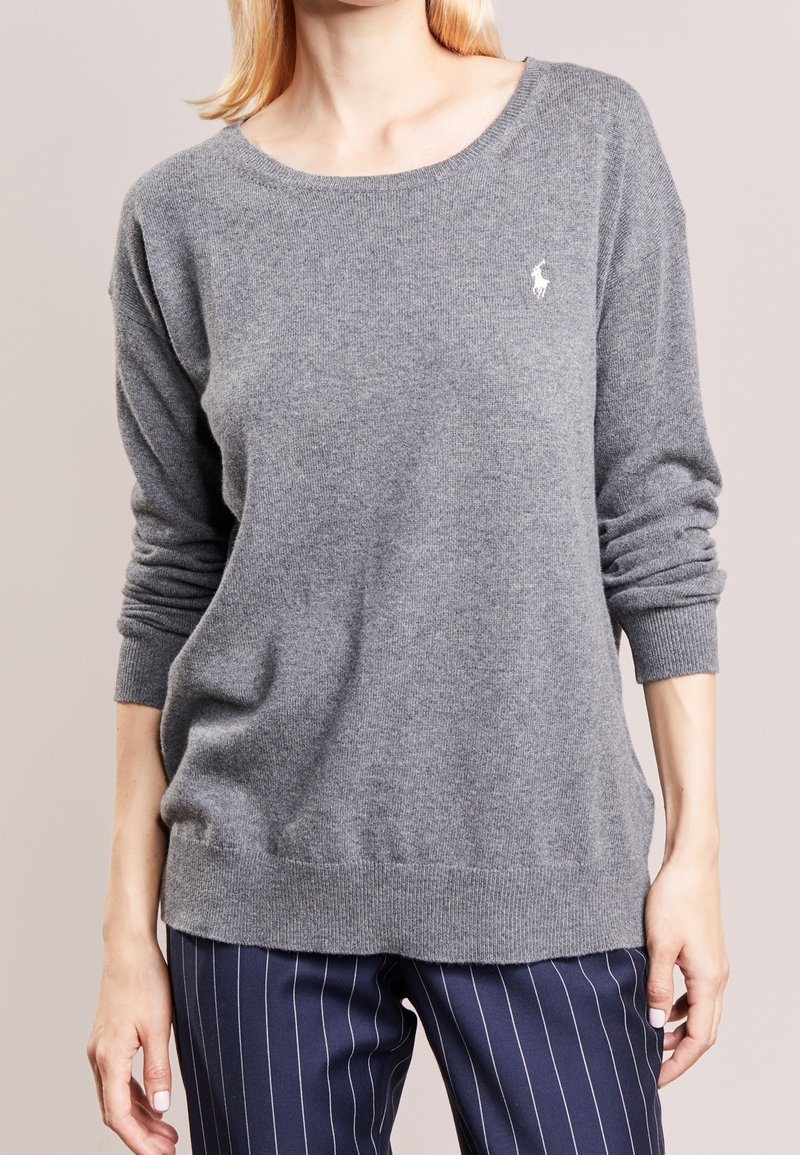 Femme portant un pull à manches longues gris clair avec un petit logo blanc brodé sur la poitrine et un pantalon bleu marine à fines rayures.