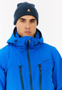 Modrá nepremokavá bunda s nastaviteľnou kapucňou, zipsovými vreckami a ventilačnými detailmi. Nosená s tmavomodrou pletenou čiapkou.