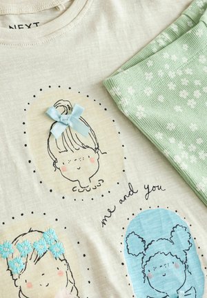 Lichtbeige T-shirt met bedrukte cartoongezichten, een blauwe strik en zwarte stippen. Gecombineerd met mintgroene leggings met witte bloemenpatronen.