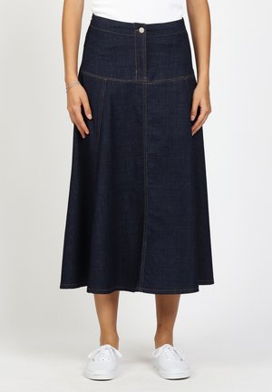 Frau trägt einen dunkelblauen A-Linien Denim-Midirock mit Knöpfen vorne und weißen Turnschuhen, stehend vor einem schlichten Hintergrund.