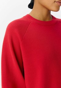 Maglione rosso con collo rotondo, maniche a raglan e una texture in maglia morbida. Il tessuto appare liscio e leggermente elasticizzato.