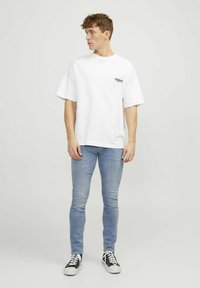 Tricou alb din bumbac cu mâneci scurte, având un mic logo negru pe piept, asortat cu blugi skinny bleumarin deschis și adidași negri.