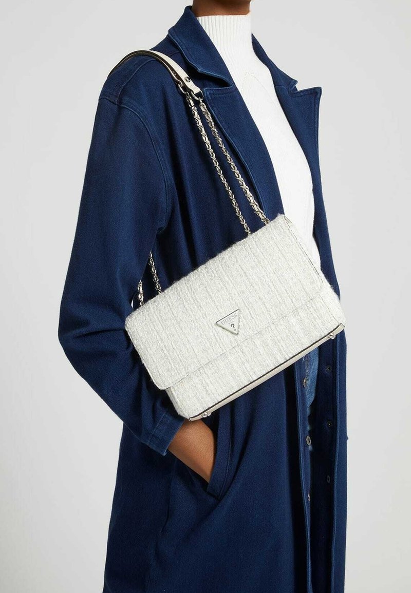 Personne en manteau bleu et col roulé blanc porte un sac à bandoulière blanc texturé avec une bandoulière en chaîne argentée et un logo triangulaire.