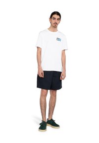 Vit bomull t-shirt med en svart och blå grafisk design på bröstet. Matchad med marinblå shorts och gröna mockaskor med beige sulor.