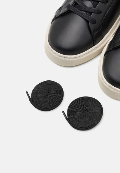 Svart skinnsneakers med en slät yta, runda vita gummisulor och svarta runda skosnören bredvid dem.