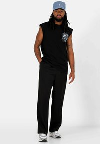 Canottiera nera senza maniche con una grafica sulla tasca anteriore, abbinata a pantaloni neri e sneakers argentate. Gli accessori includono un cappellino blu a fantasia.