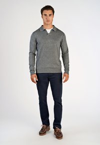 Mand står iført en grå langærmet polo sweater, mørke bukser og brune lædersko mod en ensfarvet hvid baggrund.