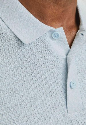 Primo piano del colletto di una polo lavorata a maglia azzurro chiaro con due bottoni, indossata da una persona con pelle di tonalità marrone media.