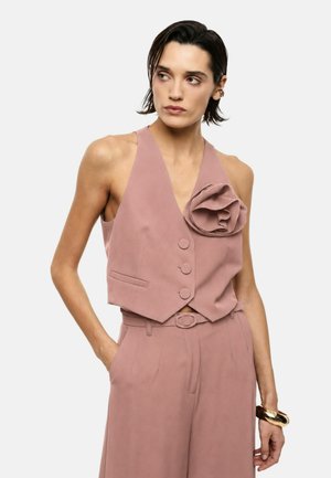 Donna che indossa un completo pantalone senza maniche rosa cipria con un grande fiore di tessuto sul gilet, in piedi con una mano in tasca.