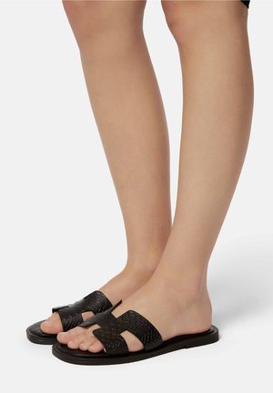 Jambes portant des sandales plates noires avec de larges brides croisées texturées, debout sur un fond clair.