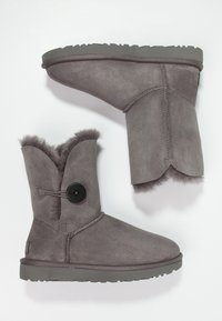 Stivali alla caviglia in suede grigio con foderatura in pelliccia, bottone laterale decorativo e suola in gomma con trazione. La parte superiore presenta un bordo ondulato.
