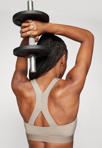 Soutien-gorge de sport beige avec un design dos nageur, présentant une texture lisse. Une personne tient une barre au-dessus de sa tête, montrant des muscles du dos toniques.