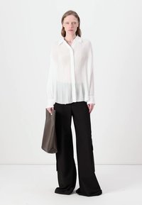 Camicia bianca a maniche lunghe plissettata con colletto, abbinata a pantaloni larghi neri. La modella tiene una borsa a tracolla grigia con texture. Sfondo neutro.