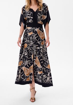 Vrouw draagt een maxi-jurk met bloemenprint in zwart, beige en bruin, met V-hals en split, in combinatie met beige hoge hakken op een witte achtergrond.