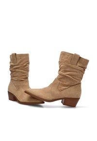 Par de botas de ante color marrón claro, hasta la mitad de la pantorrilla, de estilo slouchy, con tacones de madera bajos y tiradores, sobre un fondo blanco.