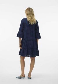 Marinblå, knälång klänning med ett lager-på-lager design och ögonlappsmönster samt trumpetärmar. Bärs med ljusblå slingback-klackar.