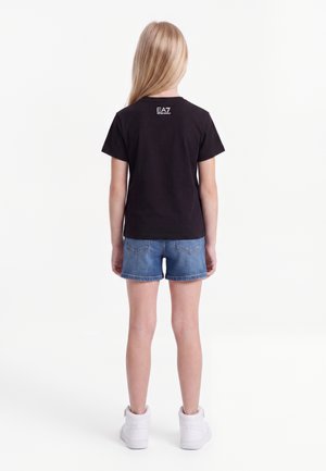 Dziewczyna o długich blond włosach, ubrana w czarny t-shirt EA7, niebieskie szorty dżinsowe i białe trampki, stojąca tyłem na białym tle.