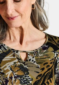 Gemusterte Bluse mit einem runden Ausschnitt und einem Schlüssellochdetail. Hergestellt aus weichem Stoff in erdigen Tönen mit floralen und abstrakten Designs.