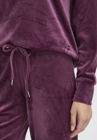 Velvet paarse loungewear set met een trekkoord in de taille en details in zilverkleur. De stof heeft een gladde, zachte textuur.