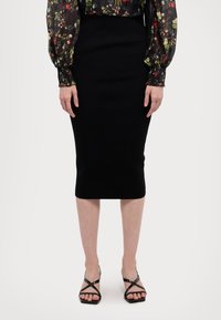 Μαύρη εφαρμοστή μολυβένια φούστα από ελαστικό ύφασμα, που φτάνει στο μέσο της γάμπας. Συνδυασμένη με μαύρα σандάλια με λουριά. Λείο υλικό, χωρίς ορατά σχέδια.