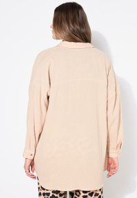 Camicia oversized beige in tessuto texturizzato, con colletto a punta, maniche lunghe e polsini con bottoni. L'orlo è leggermente curvato sul retro.