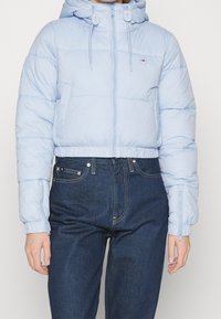 Osoba v světlemodré zkrácené péřové bundě s kapucí a tmavě modrých džínách s vysokým pasem na jednoduchém pozadí.