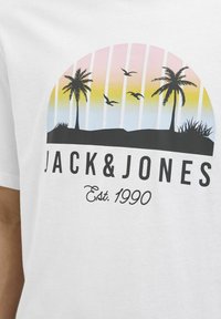 Bílé tričko s grafickým designem, zahrnujícím gradient západu slunce, palmy, ptáky a text "JACK & JONES Est. 1990" v černé barvě.