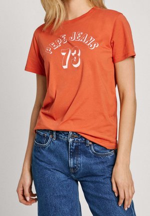 T-shirt en coton orange à manches courtes, avec les inscriptions "PEPE JEANS" et "73" en lettres blanches sur le devant. Porté avec un jean en denim bleu.