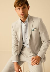 Costume gris clair en tissu texturé, avec une chemise à motifs et un pochette en bleu à fleurs. Coupe slim avec des accents simples.