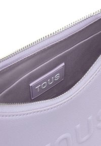 Borsetta in pelle lavanda con ferramenta in argento. Presenta il logo "TOUS" in rilievo e una fodera interna liscia. Chiusura con cerniera lungo la parte superiore.