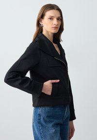 Zwarte cropped jack met brede kraag, rits aan de voorkant en zijzakken. Soepele stof en minimale naden. Gecombineerd met lichtblauwe denim jeans.