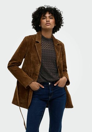 Femme aux cheveux bouclés portant une veste en daim marron, un haut noir à motifs et un jean bleu, debout les mains dans les poches devant un fond uni.