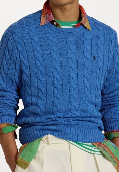 Royal blue cable-knit sweater with ribbed cuffs and hem. Worn over a multicolored plaid shirt with green and white stripes.