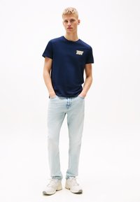 Marine bomulls t-skjorte med en liten kremfarget logo på brystet, kombinert med lyseblå denimjeans og hvite joggesko. Enkelt design.