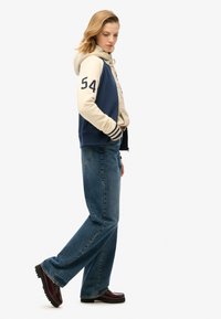 Chaqueta varsity azul marino y crema con el número 54, superpuesta sobre una sudadera ligera con capucha. Combinada con jeans acampanados azules y mocasines burdeos.