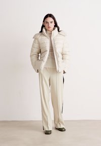 Casaco puffer bege com colarinho estruturado, fecho de zip e bolsos laterais, combinado com calças crème com riscas laterais pretas.
