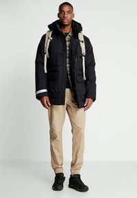 Czarna kurtka puchowa z kapturem, z przednimi kieszeniami i odblaskowymi akcentami. Nosi ją na koszuli w kratę, w połączeniu z beżowymi spodniami i czarnymi butami.