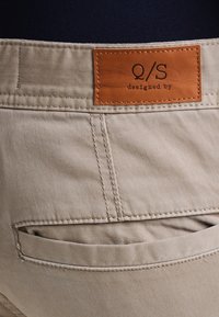 Pantalon en coton beige avec une étiquette en cuir portant la mention "Q/S designed by". Présente des coutures visibles et une poche arrière.