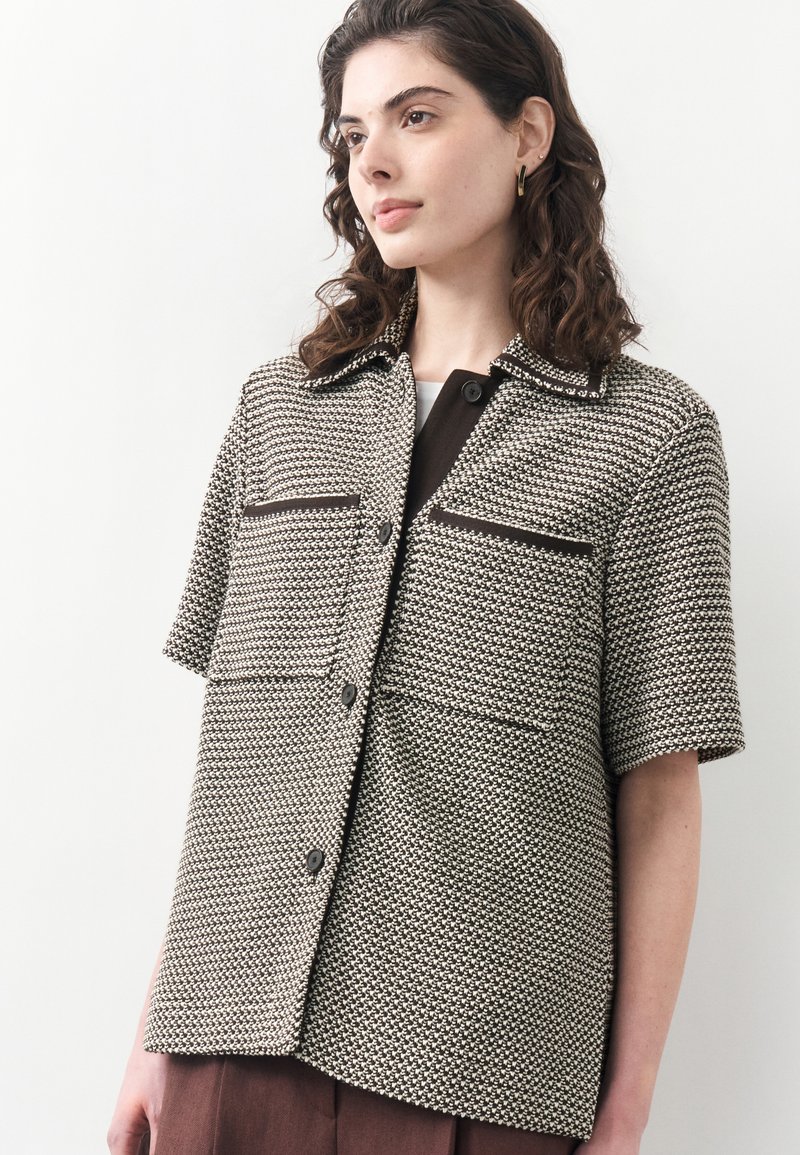 Jeune femme aux cheveux bruns bouclés portant une chemise à manches courtes noire et blanche à motifs avec des poches devant.