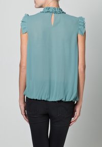 Blusa teal realizzata in tessuto leggero, con colletto arricciato e maniche a sbuffo, dotata di una scollatura a chiave sul retro e orlo gathered.