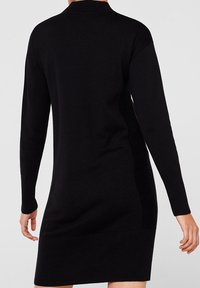 Robe pull tricotée noire avec des manches longues, un col haut, un ourlet côtelé et des détails sur les côtés, présentant une texture lisse et une silhouette ajustée.