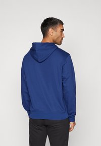 Sweatshirt bleu à capuche avec une coupe décontractée. Il présente des poignets et un ourlet côtelés, une texture douce et une poche kangourou. Vue arrière affichée.