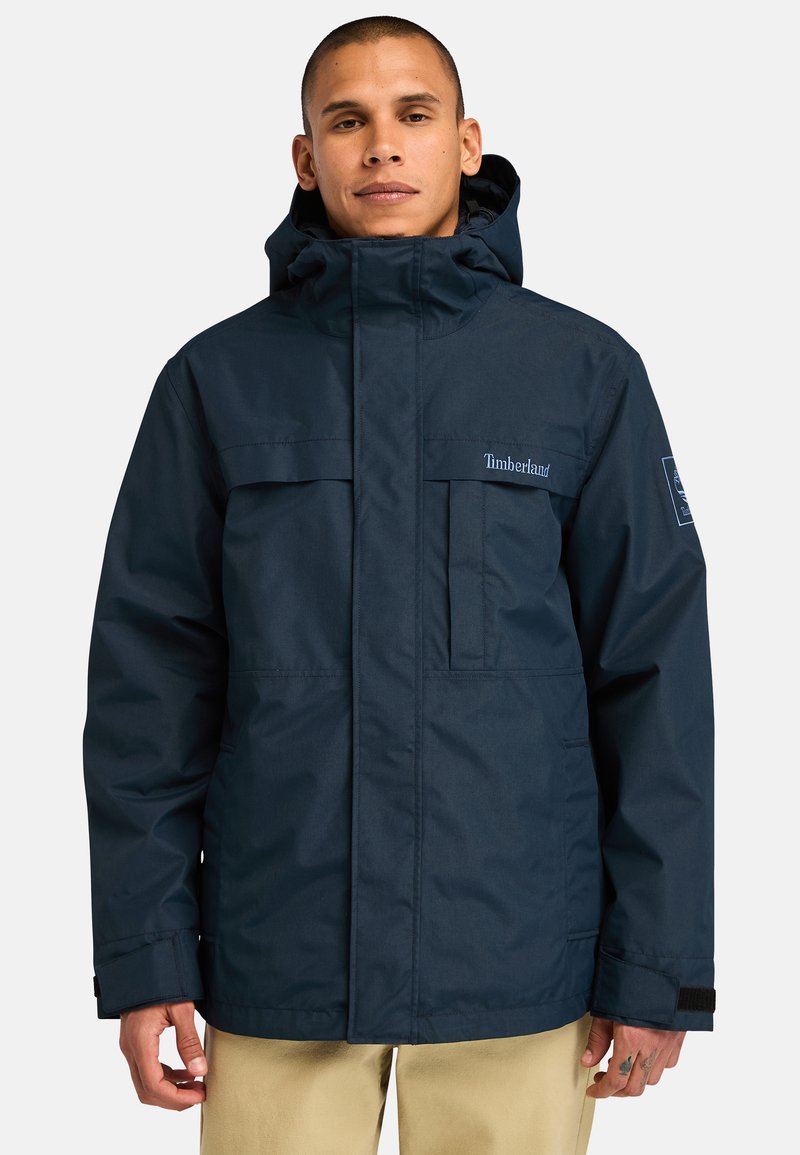 Navy waterdichte jas met capuchon, ritssluiting aan de voorkant, twee borstzakken en verstelbare mouwbanden. Getextureerde stof en logo-accent.