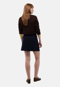 Pull marron avec des poignets jaunes, associé à une jupe bleu marine. Cheveux mi-longs, chaussures marron, vu de dos. Texture lisse, design décontracté.