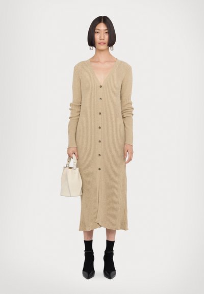 Beige geribbelde gebreide cardigan jurk met lange mouwen en knoopsluiting. Elegante V-hals, gecombineerd met een witte handtas en zwarte enkellaarsjes.