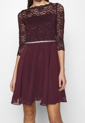 Femme portant une robe bordeaux avec un haut en dentelle, manches trois-quarts, une ceinture à paillettes argentées, et une jupe fluide en chiffon.