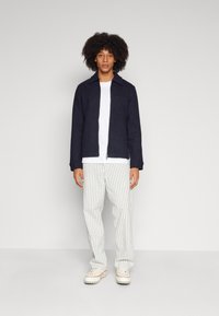 Navy zip-up jacket with a collar and pocket, paired with white striped trousers and beige sneakers. Smooth texture throughout the outfit.