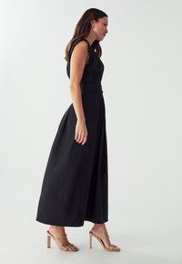 Abito maxi nero senza maniche con vita aderente e gonna fluida. Tessuto morbido, presenta una leggera lucentezza. Indossato con tacchi nudi con cinturini.