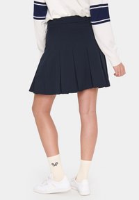Fustă scurtă plisată bleumarin cu talie înaltă, confecționată dintr-un material texturat. Purtați cu adidași albi și ciorapi până la genunchi cu un design inspirat de racheta de tenis.